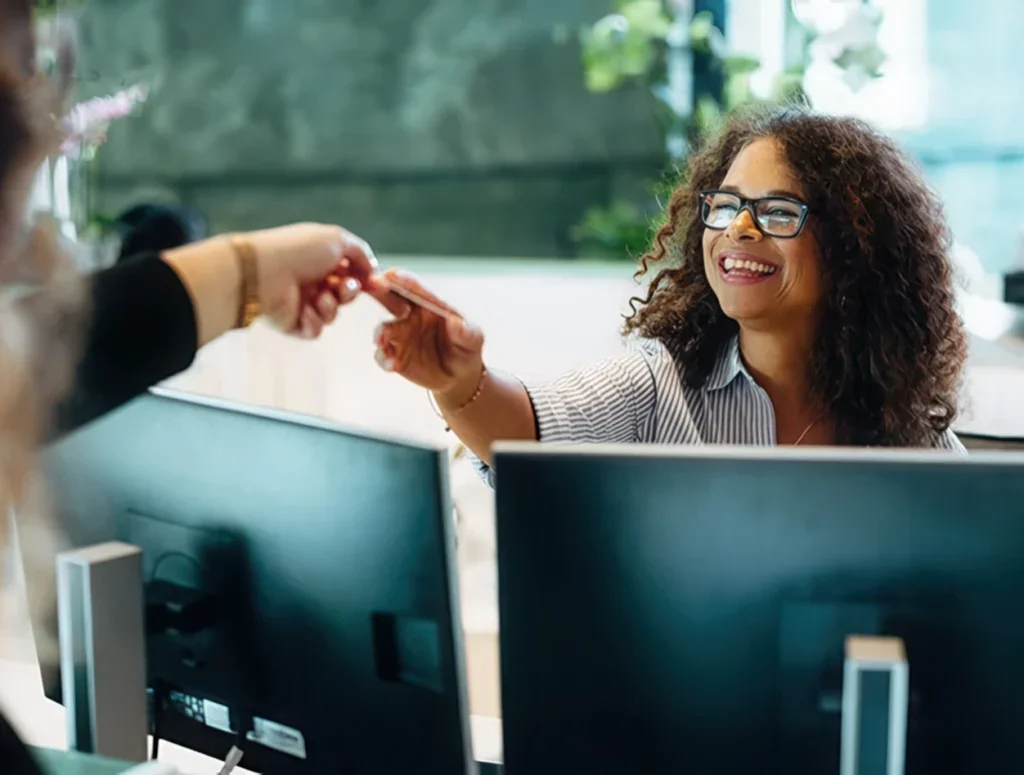 Smiling woman, interacting with another employee over a double monitor.